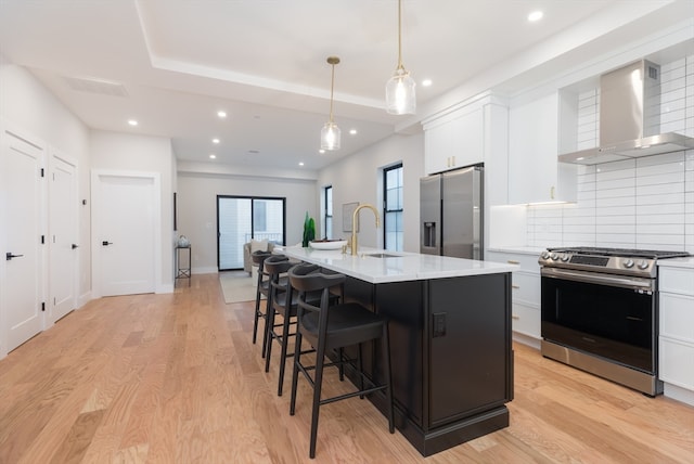 kitchen with appliances with stainless steel finishes, wall chimney exhaust hood, sink, a center island with sink, and white cabinets