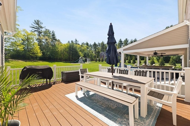 wooden terrace featuring a playground, a lawn, outdoor dining area, and a ceiling fan