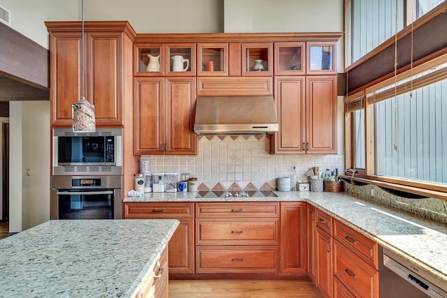 kitchen with light stone countertops, appliances with stainless steel finishes, brown cabinets, backsplash, and ventilation hood