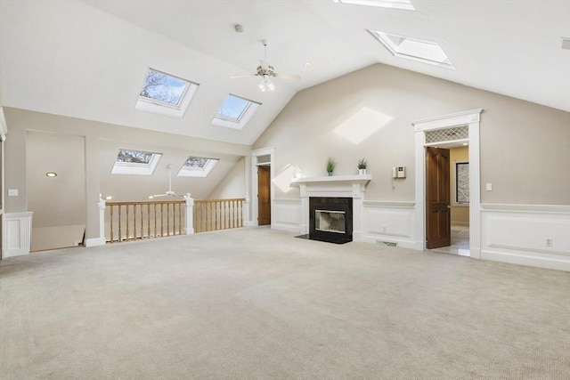 unfurnished living room featuring light colored carpet, a fireplace with flush hearth, a ceiling fan, vaulted ceiling, and wainscoting