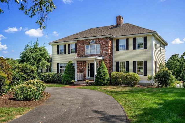 view of front of home featuring brick siding, a chimney, a shingled roof, a front yard, and driveway