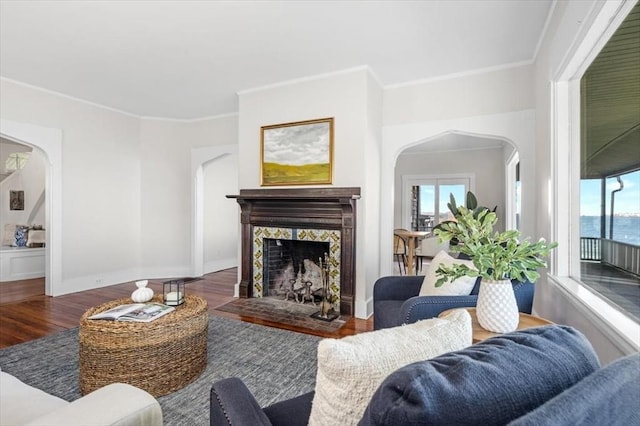 living area with arched walkways, wood finished floors, a tiled fireplace, and crown molding