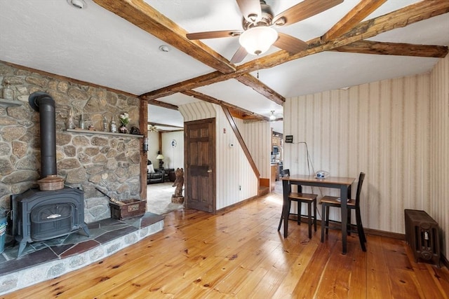 dining space with ceiling fan, hardwood / wood-style flooring, beamed ceiling, and a wood stove