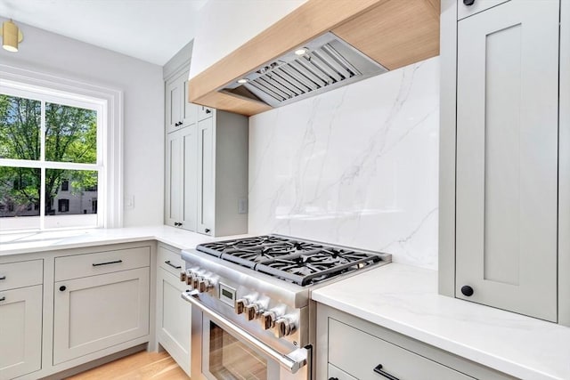 kitchen featuring decorative backsplash, high end stove, light stone countertops, premium range hood, and light wood-style floors