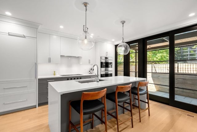 kitchen with decorative light fixtures, white cabinetry, sink, stainless steel double oven, and a center island with sink