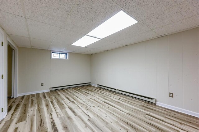 empty room with a baseboard radiator, a drop ceiling, light wood finished floors, and baseboards