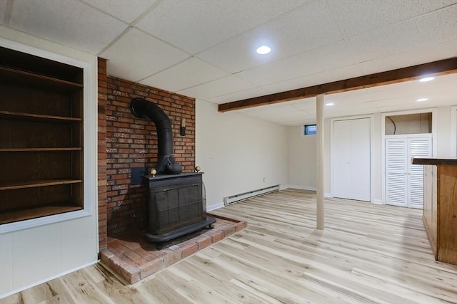 unfurnished living room with a baseboard heating unit, wood finished floors, a drop ceiling, a wood stove, and baseboards
