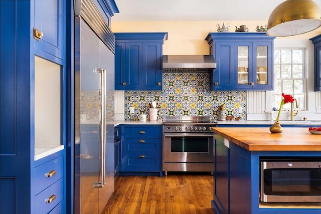 kitchen with blue cabinets, high end appliances, butcher block counters, glass insert cabinets, and dark wood-type flooring