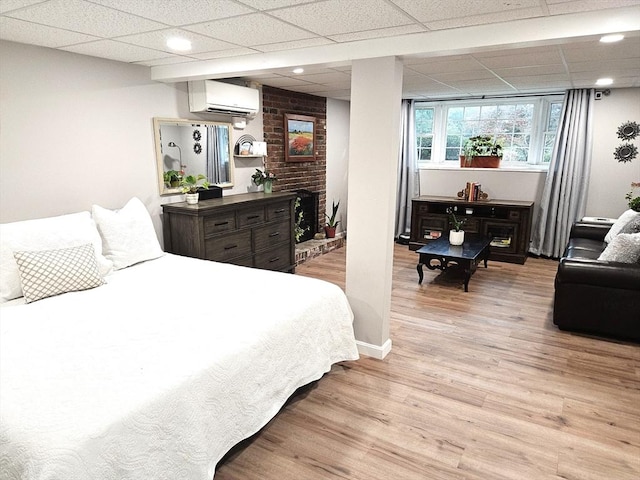 bedroom with a wall mounted air conditioner, light wood-style flooring, a paneled ceiling, baseboards, and a brick fireplace