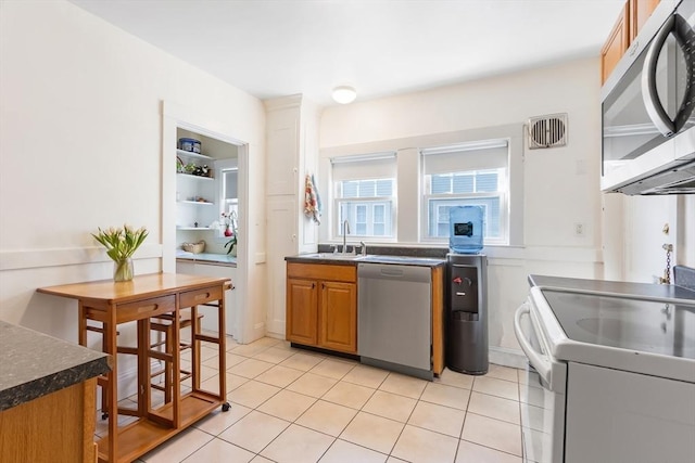 kitchen with brown cabinets, stainless steel appliances, dark countertops, light tile patterned flooring, and a sink