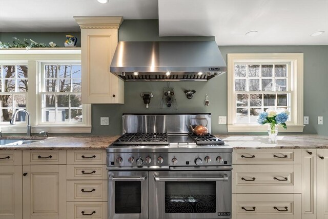 kitchen featuring wall chimney range hood, range with two ovens, light stone countertops, and a sink