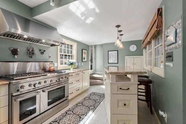 kitchen featuring a breakfast bar area, light stone countertops, range with two ovens, light tile patterned flooring, and wall chimney range hood