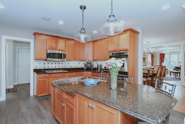 kitchen with pendant lighting, wood finished floors, appliances with stainless steel finishes, a center island with sink, and dark stone countertops