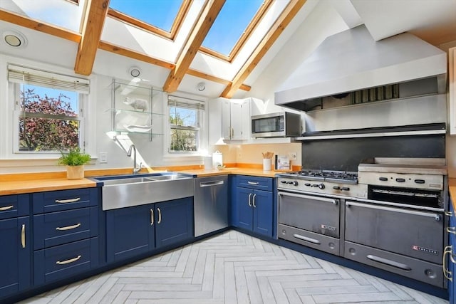 kitchen featuring wall chimney range hood, appliances with stainless steel finishes, a sink, vaulted ceiling with skylight, and blue cabinets
