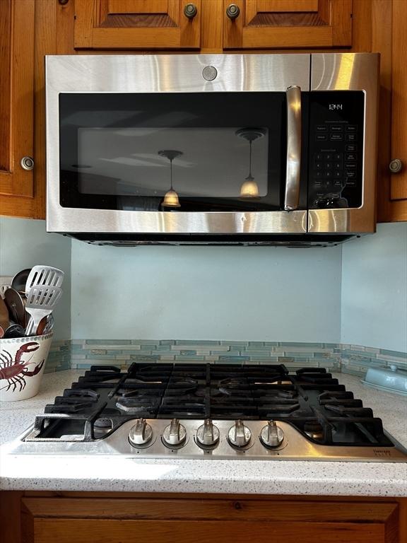 details featuring stainless steel microwave, tasteful backsplash, gas stovetop, brown cabinetry, and light stone countertops