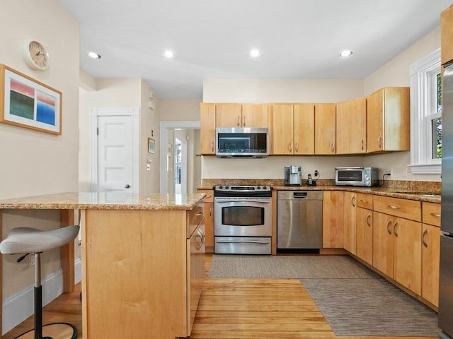 110 Woerd Ave, Waltham, MA 02453 - kitchen with appliances with stainless steel finishes, light wood-style floors, light stone countertops, light brown cabinetry, and a kitchen breakfast bar