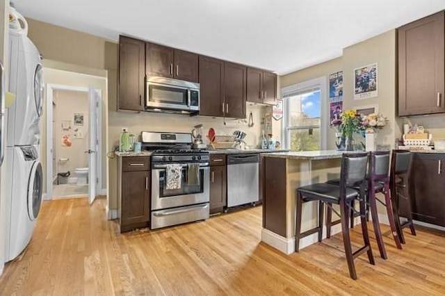 kitchen with appliances with stainless steel finishes, stacked washer and clothes dryer, dark brown cabinetry, light wood-style flooring, and a breakfast bar area