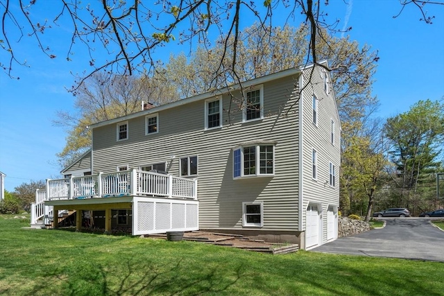 rear view of property with an attached garage, a chimney, a wooden deck, aphalt driveway, and a yard