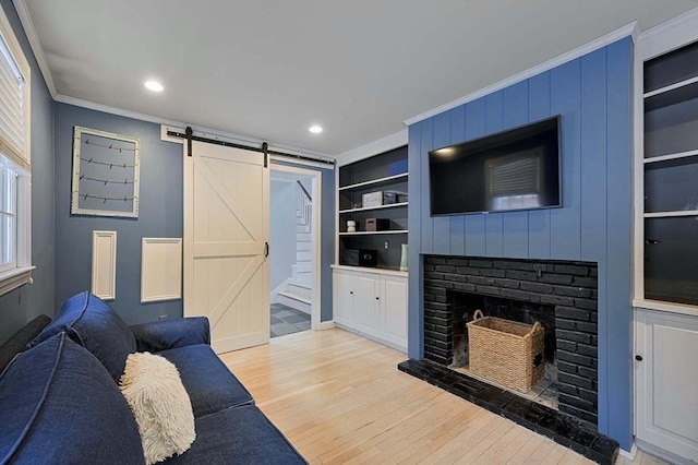 living room with a barn door, built in features, light wood-style flooring, crown molding, and a brick fireplace