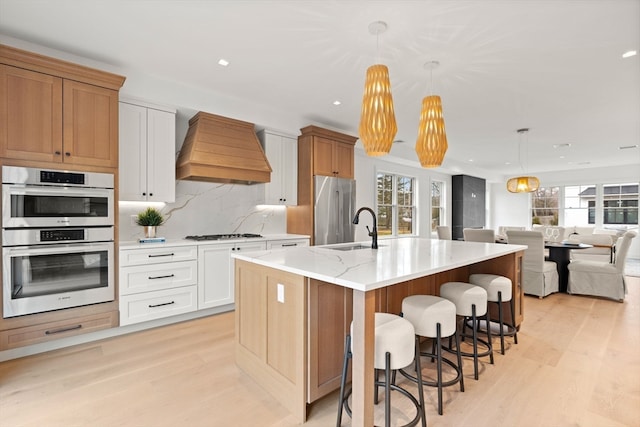 kitchen with premium range hood, an island with sink, pendant lighting, white cabinets, and appliances with stainless steel finishes