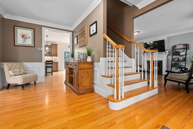 staircase with a decorative wall, wood finished floors, ornamental molding, and a warm lit fireplace