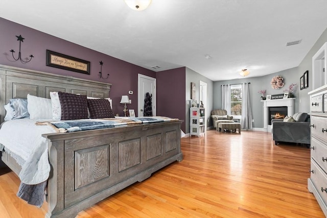 bedroom featuring light wood finished floors, a warm lit fireplace, baseboards, and visible vents