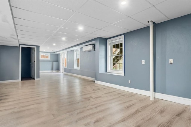 basement with a drop ceiling, wood finished floors, baseboards, and a wall unit AC