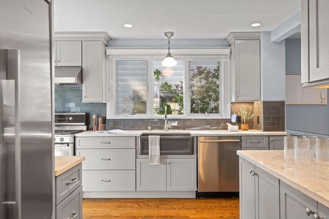 kitchen featuring wall chimney range hood, appliances with stainless steel finishes, hanging light fixtures, a sink, and gray cabinetry