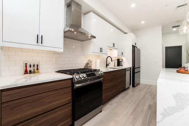 kitchen featuring light wood finished floors, white cabinets, wall chimney exhaust hood, stainless steel appliances, and a sink