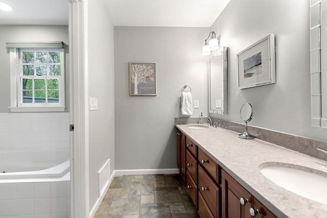 full bath featuring baseboards, double vanity, a garden tub, and stone finish flooring