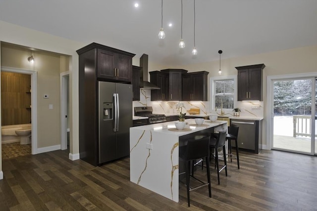 kitchen with appliances with stainless steel finishes, decorative backsplash, a kitchen island, decorative light fixtures, and wall chimney exhaust hood