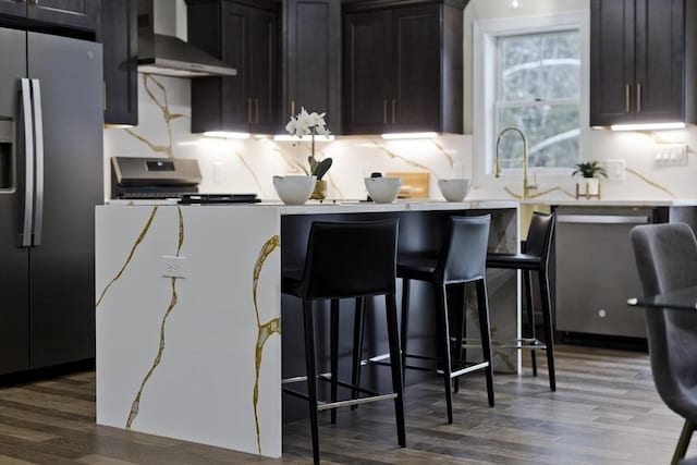 kitchen with hardwood / wood-style floors, appliances with stainless steel finishes, dark brown cabinetry, wall chimney exhaust hood, and a kitchen bar