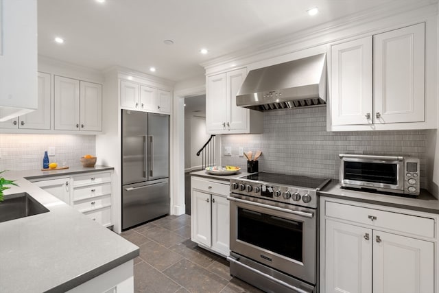 kitchen with wall chimney range hood, dark tile floors, backsplash, premium appliances, and white cabinets