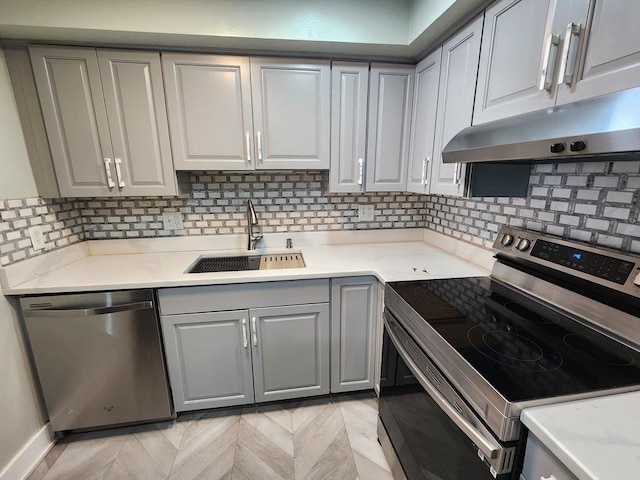 kitchen with gray cabinets, a sink, under cabinet range hood, backsplash, and stainless steel appliances