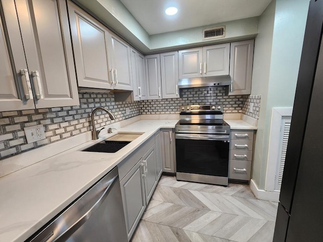 kitchen with gray cabinets, a sink, under cabinet range hood, tasteful backsplash, and appliances with stainless steel finishes