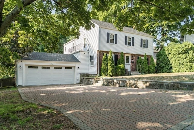 colonial inspired home with decorative driveway, brick siding, a balcony, and roof with shingles