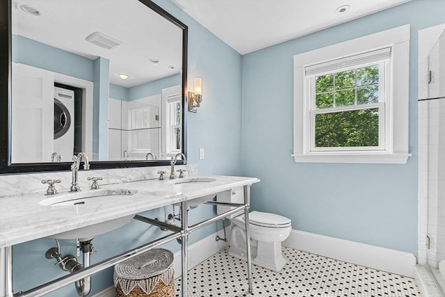 bathroom with estacked washer and dryer, toilet, baseboards, tiled shower, and double vanity