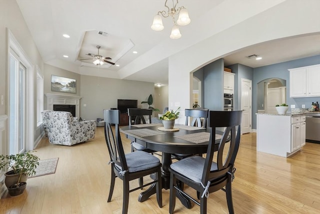 dining space featuring light wood finished floors, a fireplace, arched walkways, ceiling fan with notable chandelier, and a tray ceiling
