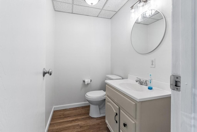 half bathroom featuring baseboards, toilet, wood finished floors, vanity, and a paneled ceiling