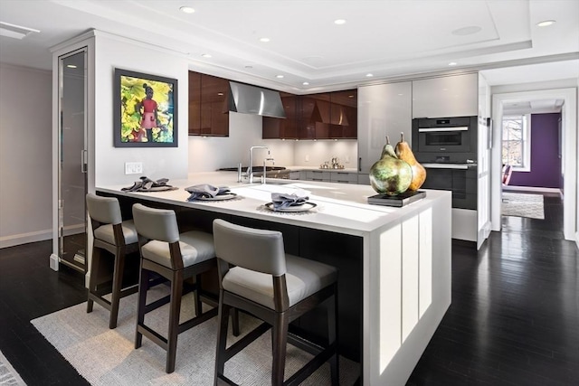 kitchen featuring a kitchen breakfast bar, sink, kitchen peninsula, wall chimney exhaust hood, and dark brown cabinetry