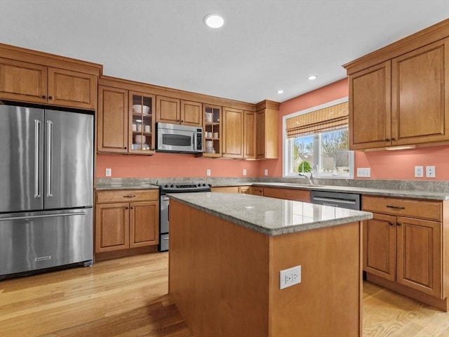 kitchen featuring appliances with stainless steel finishes, brown cabinetry, light wood-type flooring, a kitchen island, and glass insert cabinets