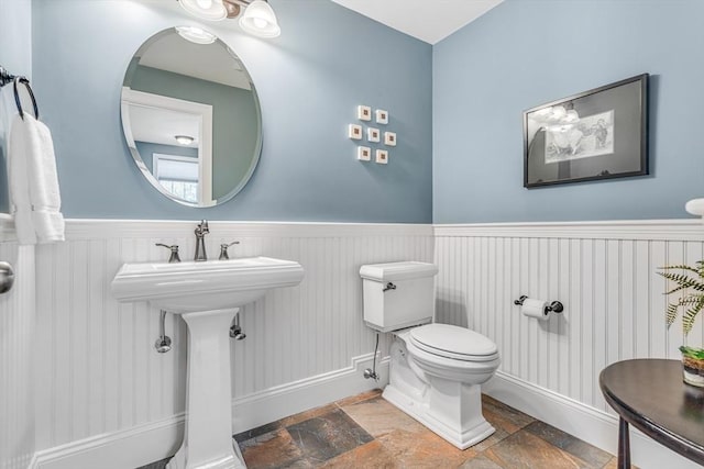 half bath featuring a sink, toilet, a wainscoted wall, stone tile floors, and baseboards