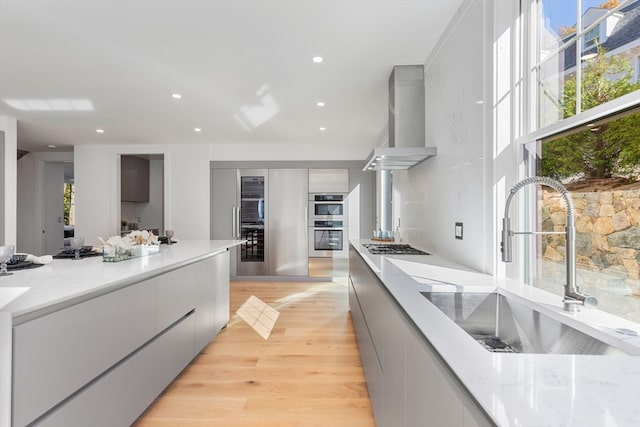 kitchen featuring stainless steel appliances, sink, wall chimney range hood, light stone countertops, and light wood-type flooring