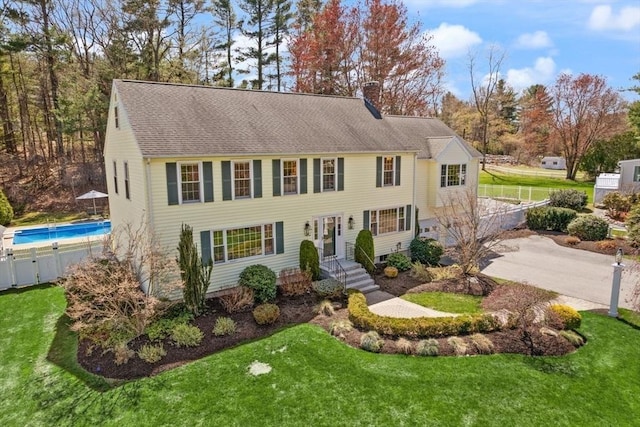 colonial-style house featuring a chimney, roof with shingles, an outdoor pool, fence, and a front lawn