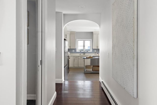 hall with a baseboard heating unit, baseboards, dark wood-style flooring, arched walkways, and a sink