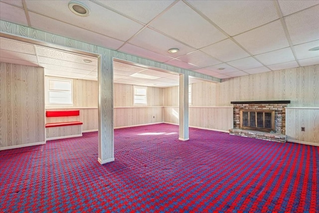 basement featuring a brick fireplace, carpet floors, a paneled ceiling, and wood walls