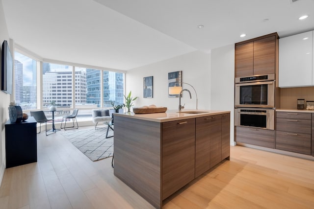 kitchen featuring light wood-type flooring, a wall of windows, stainless steel double oven, sink, and a center island with sink