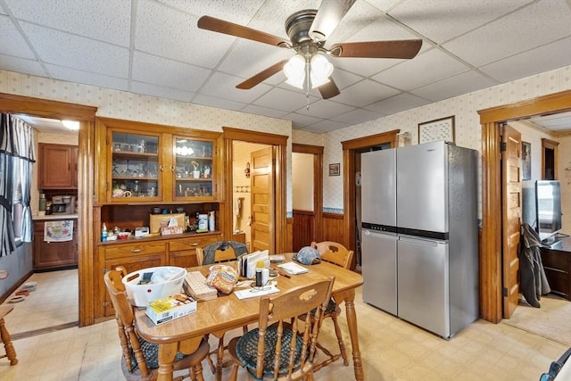 dining space with wallpapered walls, a wainscoted wall, light flooring, ceiling fan, and a paneled ceiling