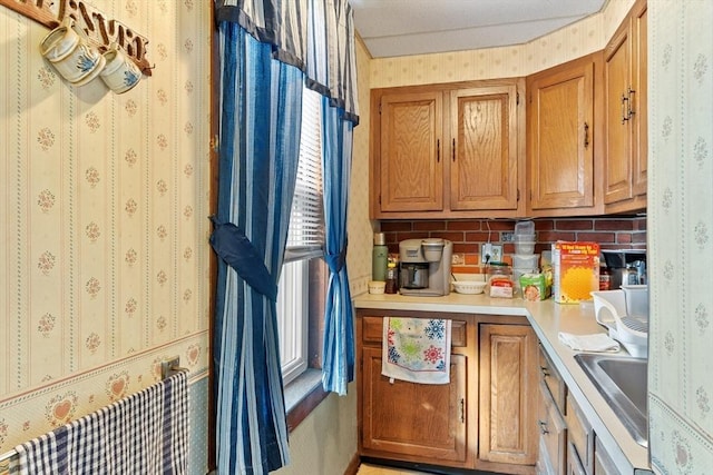 kitchen with light countertops, wallpapered walls, backsplash, brown cabinetry, and a sink