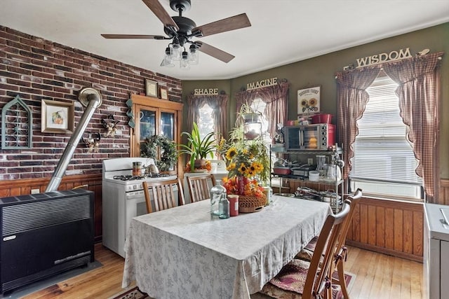 dining space with a wood stove, light wood-style flooring, ceiling fan, and brick wall